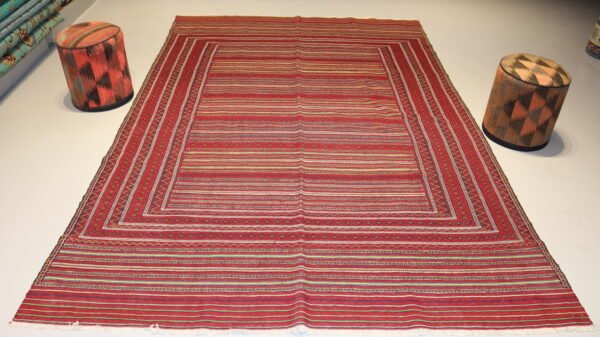 Vintage handwoven kilim rug featuring striped geometric patterns in red, black, and earthy tones, displayed in a modern showroom with matching patterned stools.